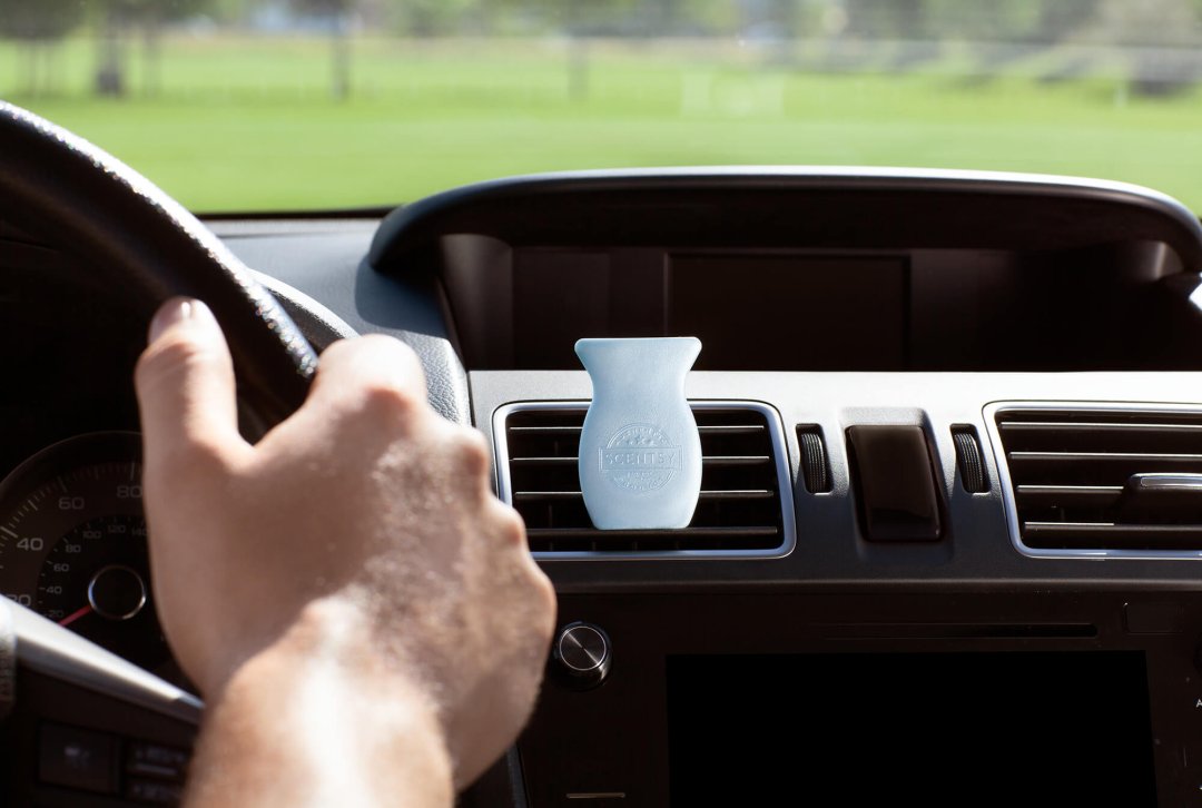 Person driving with hands on the wheel and a Car Bar Clip plugged into the vent beside him scented in Clothesline