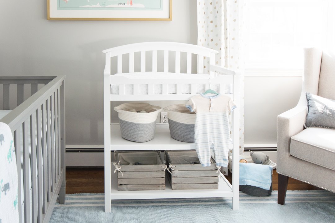 A white changing table with grey baskets for storage below, near a grey crib