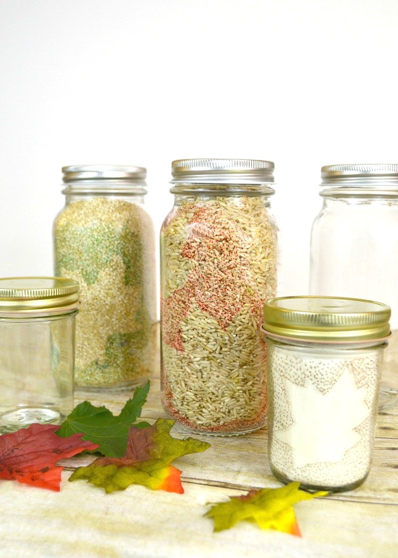 3 jars filled with pantry staples decorated with leaf motifs and 2 plain jars on wood table with leaves