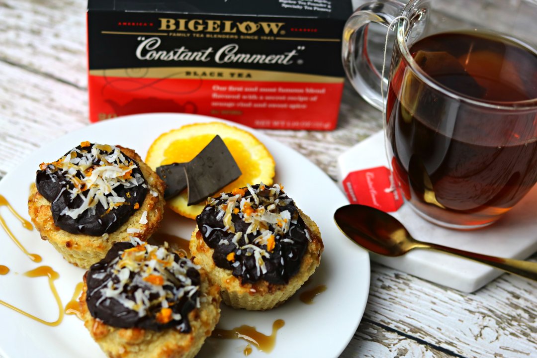 A plate of Keto Orange Muffins with orange chocolate glaze next to a glass of tea, and in front of a box of Bigelow Constant Comment