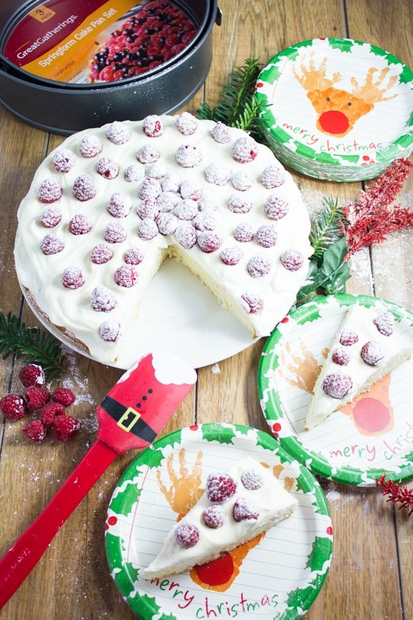 overhead of festive Vanilla Whipped Cream and Raspberry Holiday Cake with a cake tin and some Christmas plates in the background